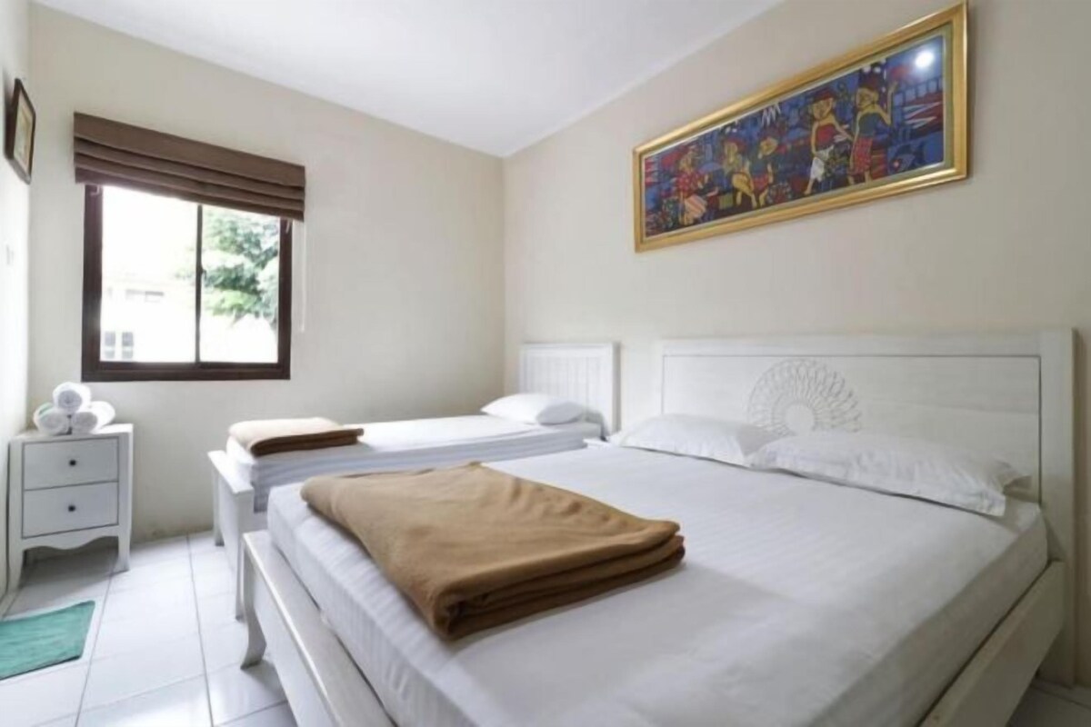 A hotel room with two beds, a window with blinds, a bedside table with a lamp, and a painting on the wall.