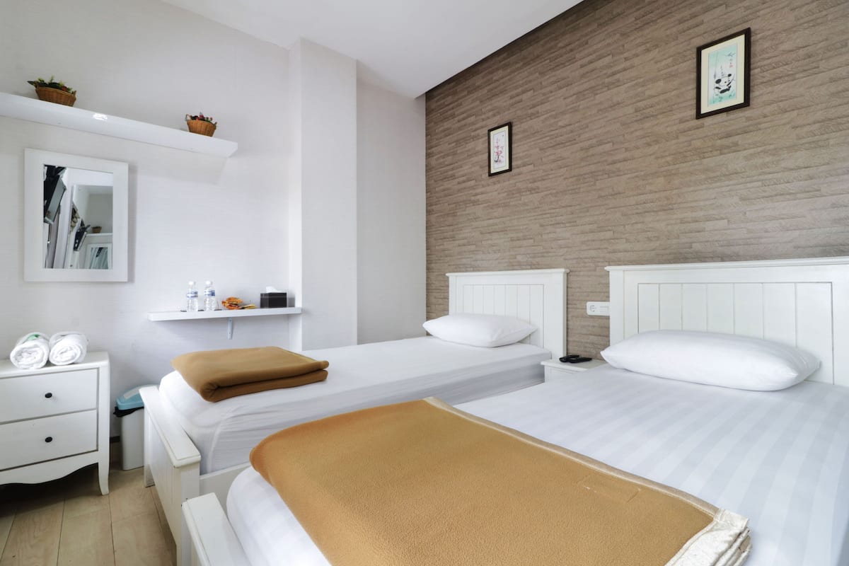 A hotel room with two beds, a white nightstand, a mirror, a shelf with toiletries, and a brick-patterned wall.