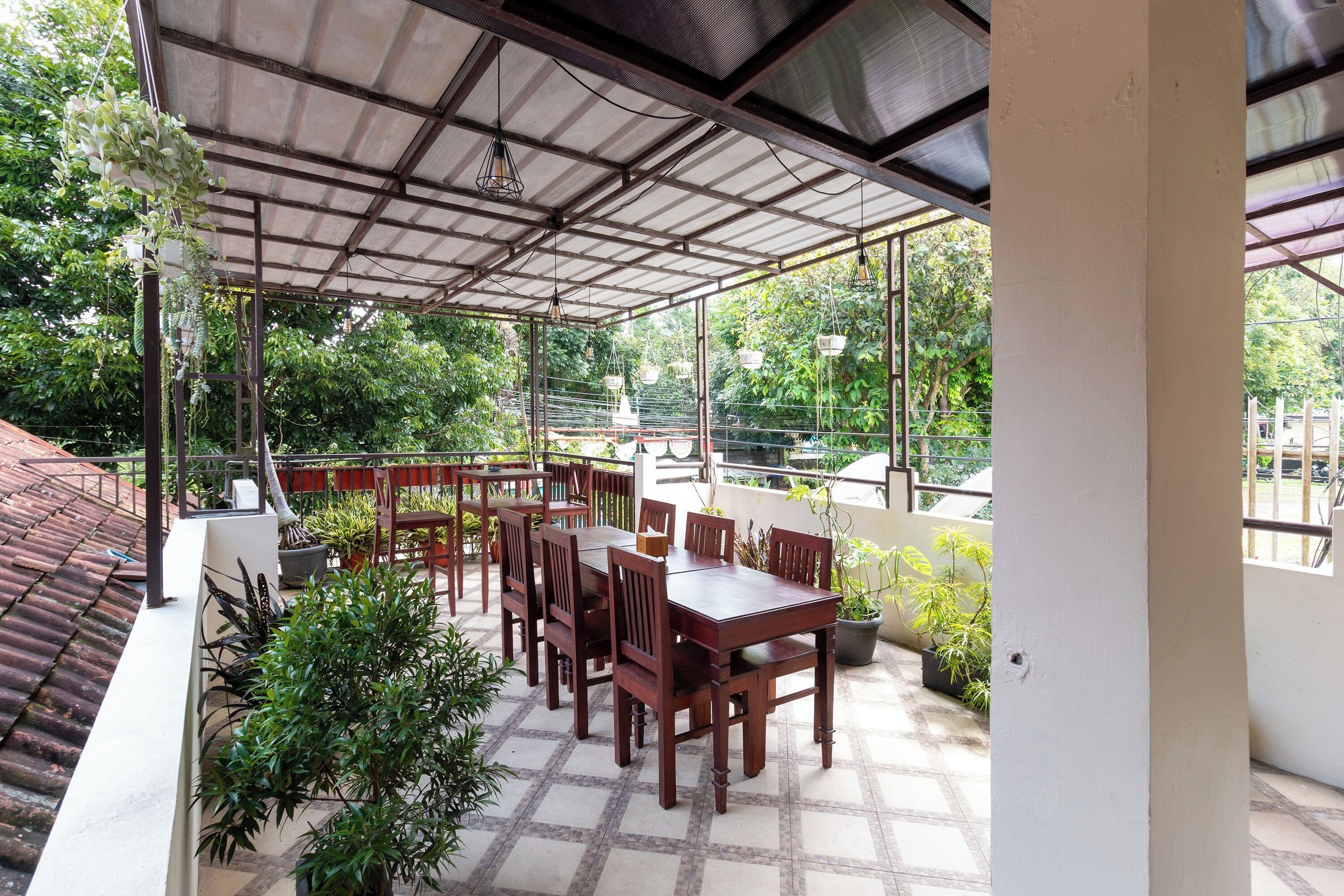 A covered outdoor dining area with a table and chairs, surrounded by potted plants and a view of greenery.