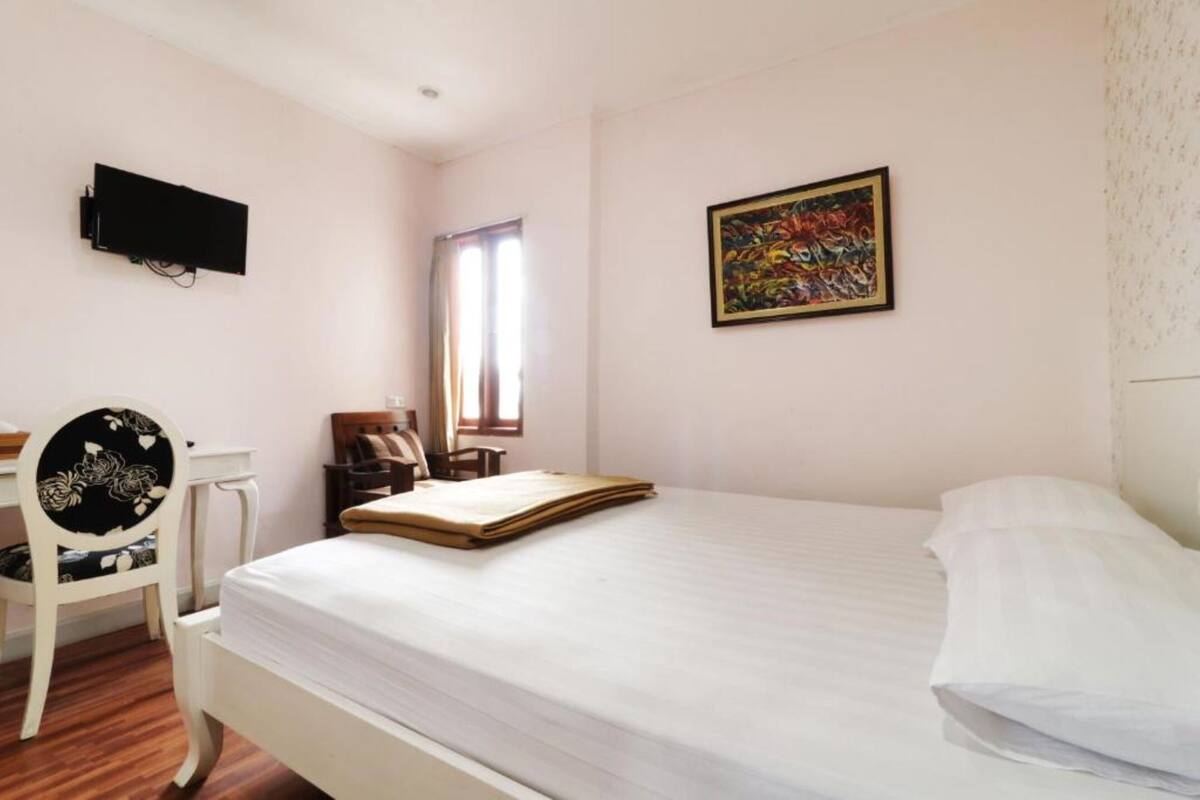 A bedroom with a bed, a desk, a chair, a television, a window, and a painting on the wall.