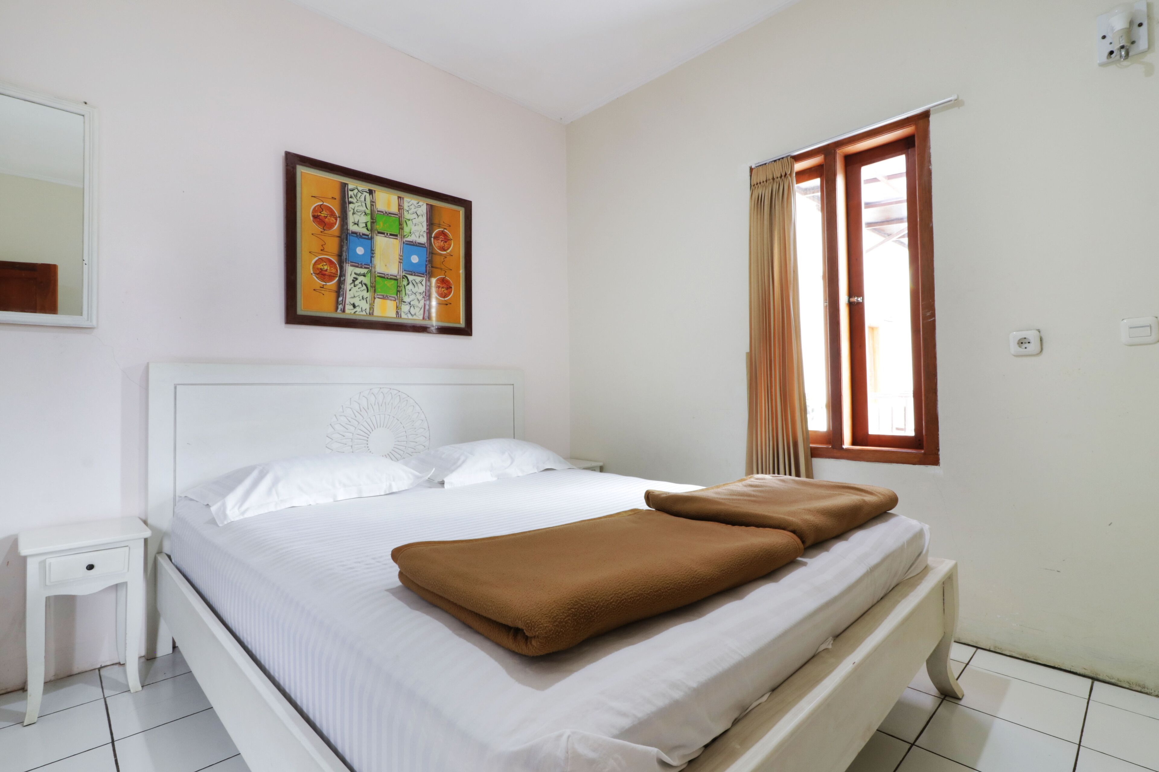A neatly made bed with white and brown bedding, a small bedside table, a colorful abstract painting on the wall, and a window with curtains.