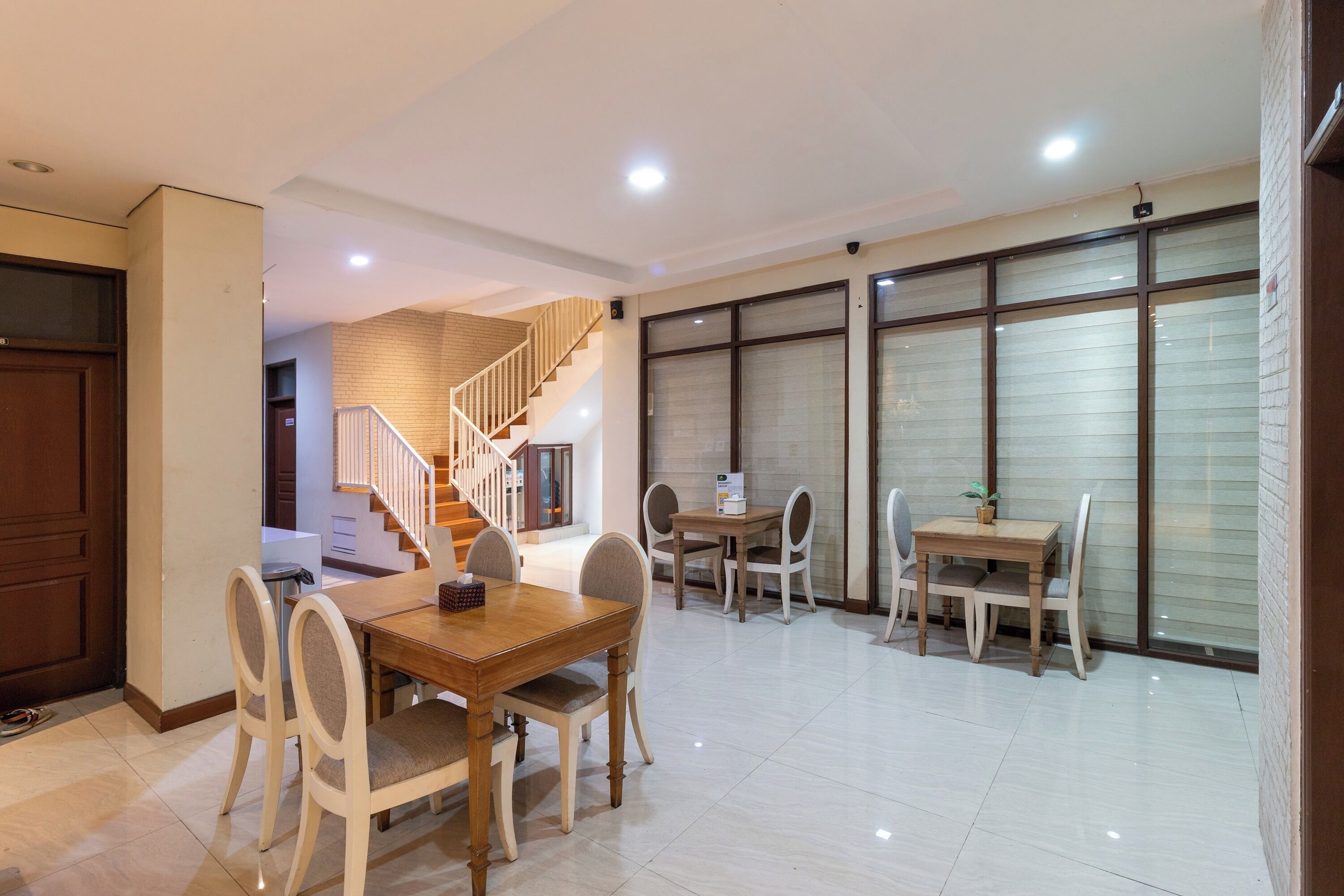 A spacious dining area with a wooden table and chairs, a staircase leading to an upper floor, and large windows with blinds.