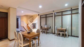 A spacious dining area with a wooden table and chairs, a staircase leading to an upper floor, and large windows with blinds.