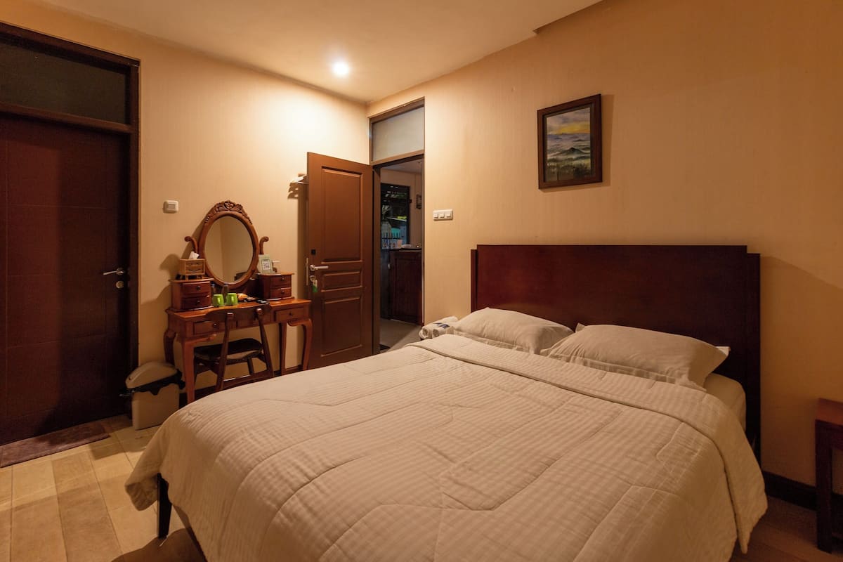 A bedroom with a large bed, a wooden dresser, a mirror, and a door leading to another room.