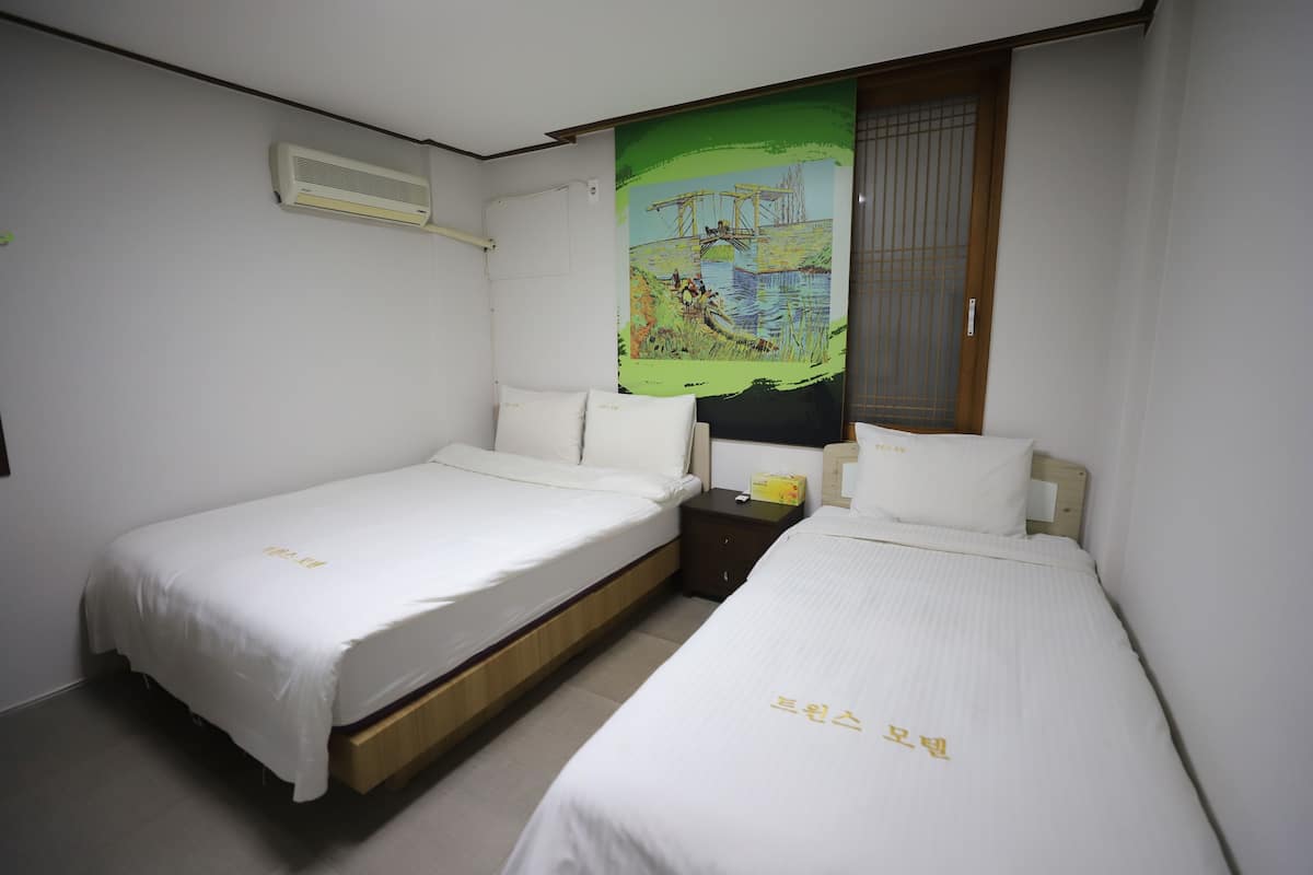 Habitación de hotel con dos camas individuales, una mesita de noche, un equipo de aire acondicionado y un cuadro grande en la pared.