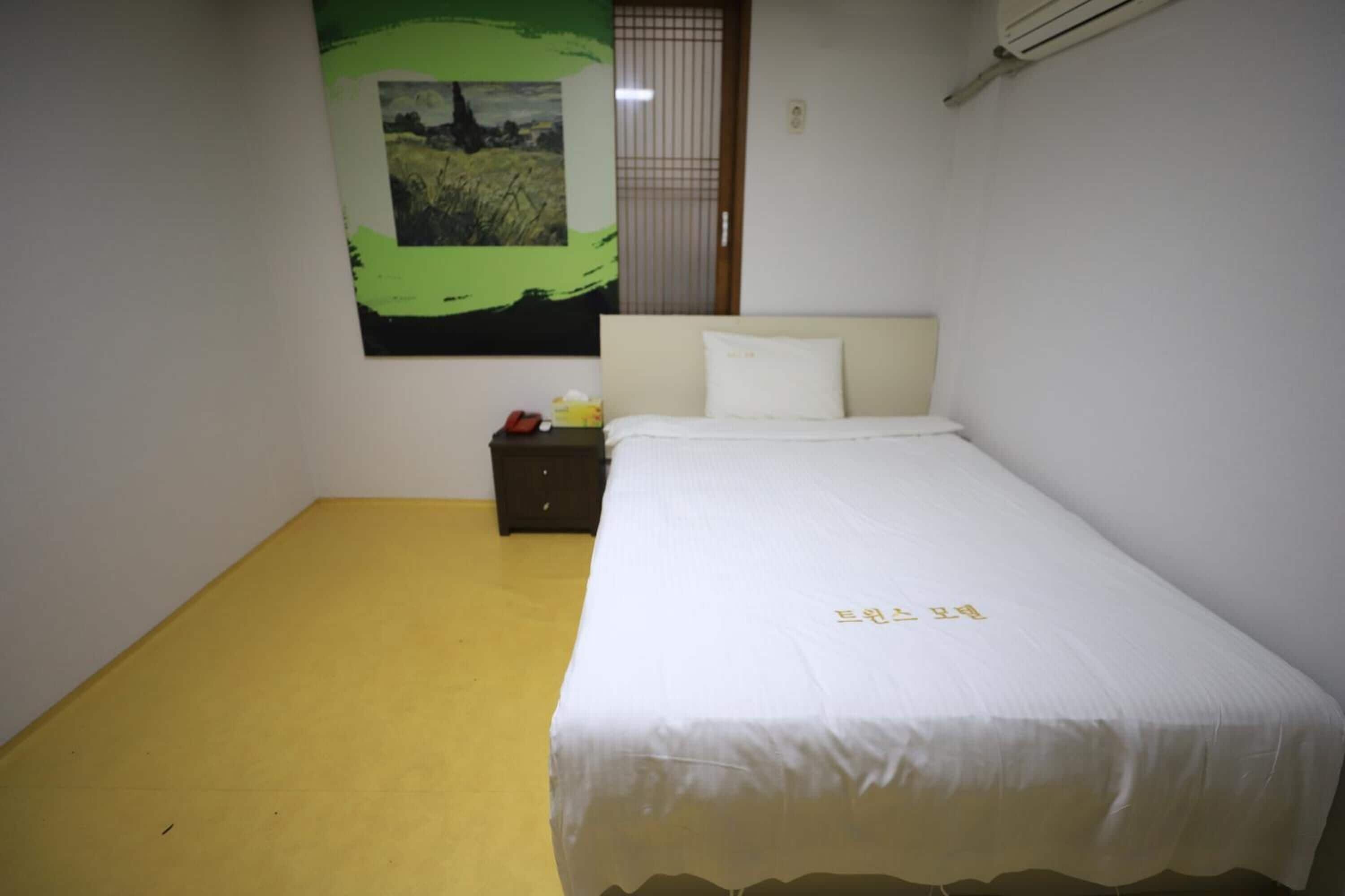 A hotel room with a bed, a nightstand, a yellow floor, a white wall, and a green painting.