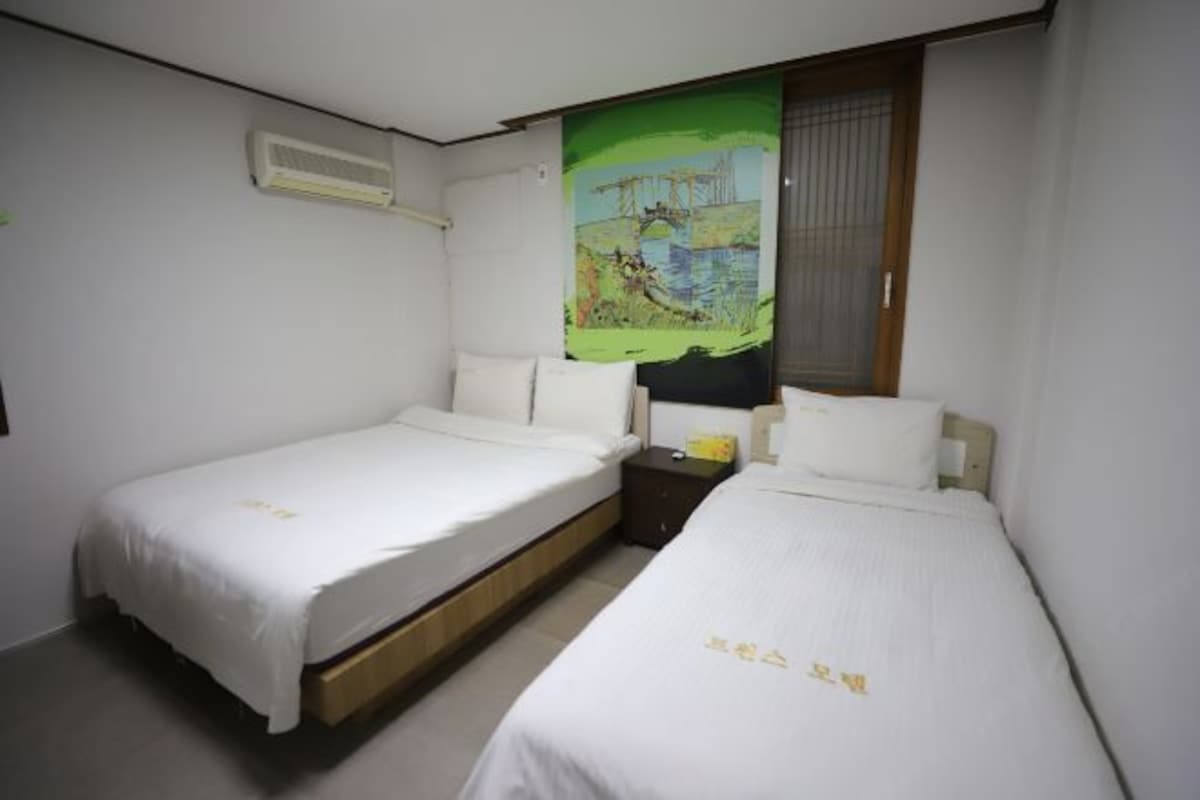 A hotel room with two single beds, a nightstand, a painting on the wall, and a window with blinds.