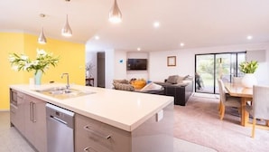 A modern kitchen with a central island, a dining area with a wooden table, and a living space with a sofa and a TV. - Euphoria House (Wynyard)