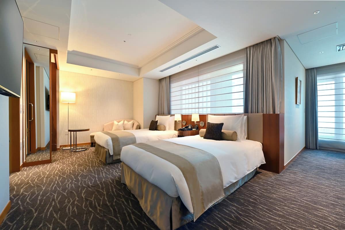 A hotel room with two beds, a sitting area, a desk, and a large window with curtains.