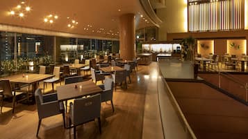 A spacious indoor dining area with multiple tables and chairs, a central pillar, and a view of the cityscape through large windows.