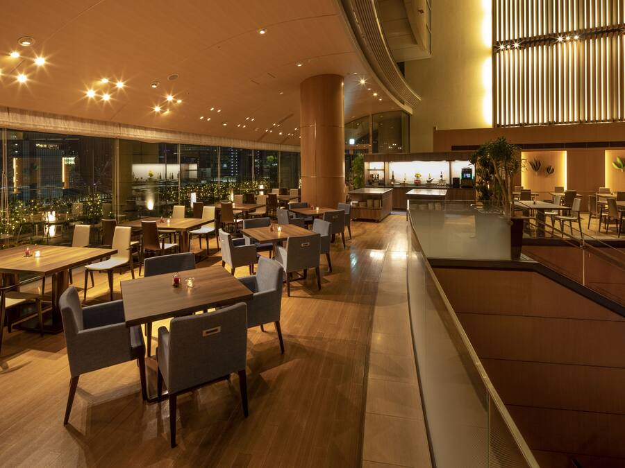 A spacious indoor dining area with multiple tables and chairs, a central pillar, and a view of the cityscape through large windows.