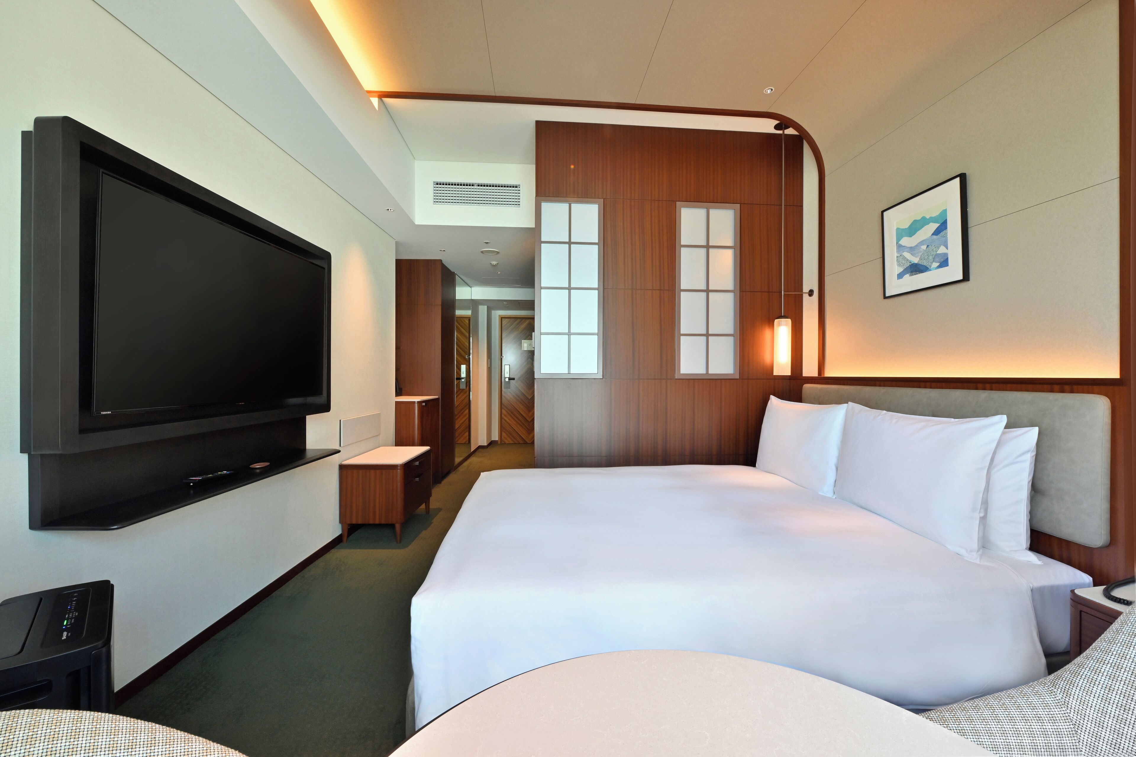 Una habitación de hotel moderna con un televisor de pantalla plana grande, una cama bien hecha, una mesita de noche y un cuadro colgado en la pared.