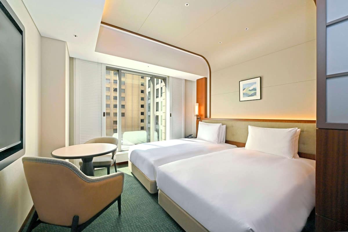 A hotel room with two beds, a small table, a chair, and a window with a view of a building.