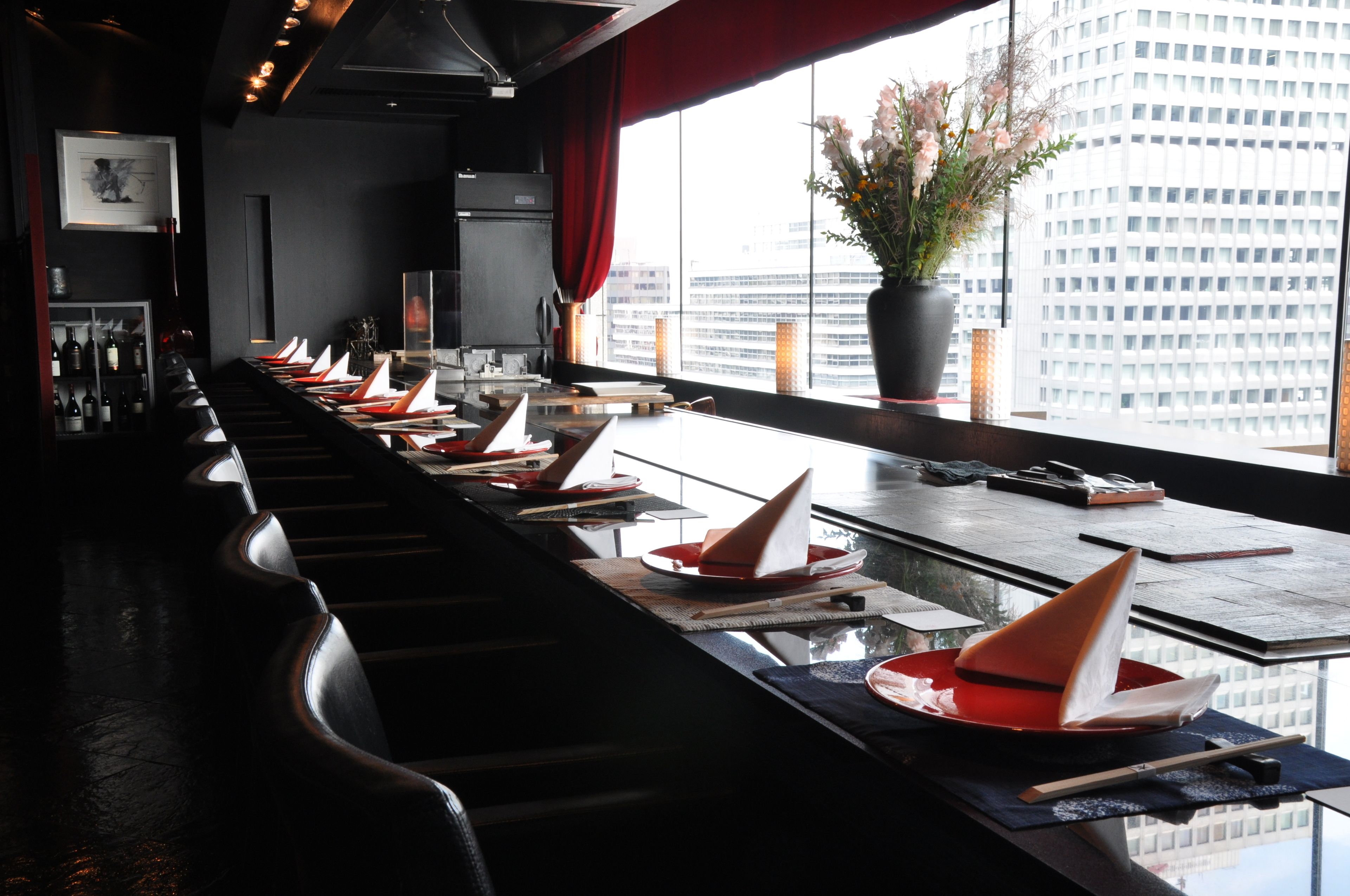 A restaurant with a long counter, multiple dining tables, and a large window with a city view.