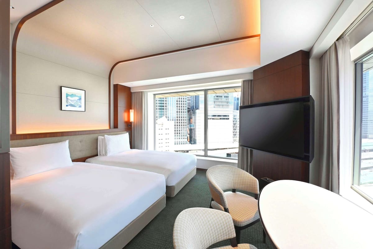 A hotel room with two beds, a flat-screen TV, a dining table, and a view of the cityscape.