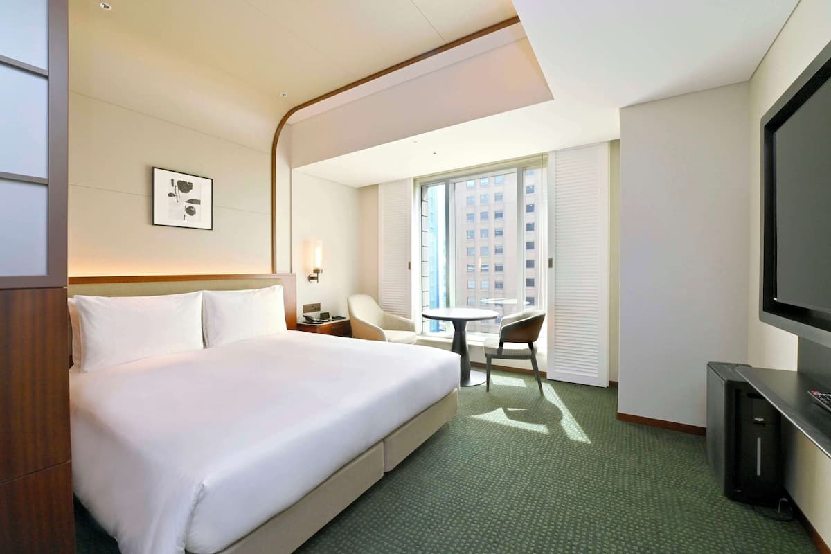 A hotel room with a large bed, a desk with a chair, a television, and a window with blinds.