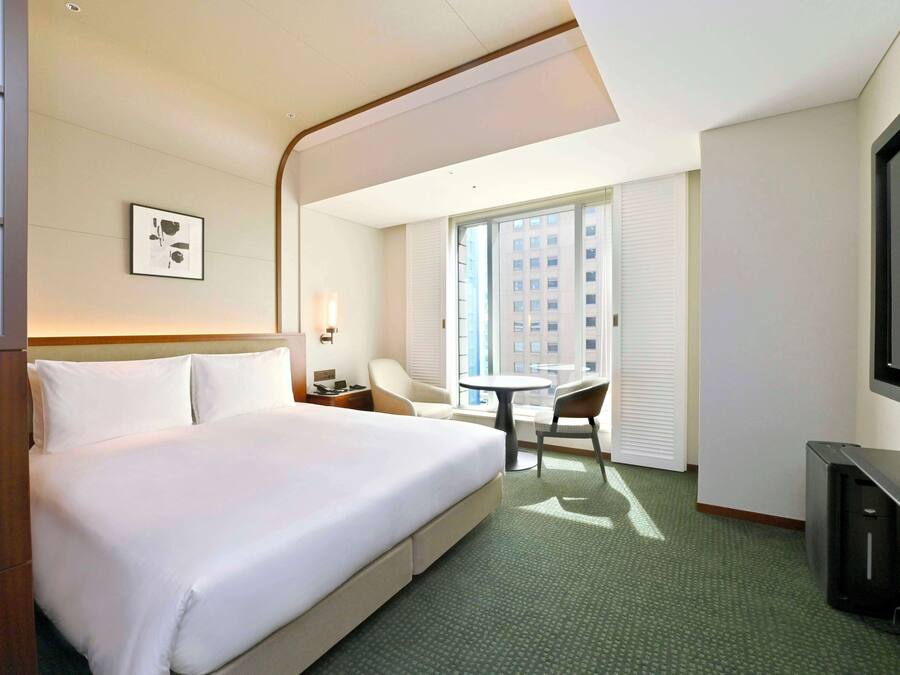 A hotel room with a large bed, a desk with a chair, a television, and a window with blinds.