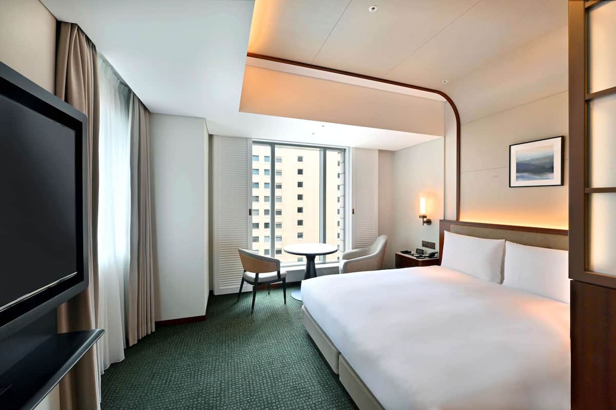 A hotel room with a large bed, a TV, a desk with a chair, a small table, and a window with a view of buildings.