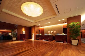 A spacious lobby with wooden flooring, a large central chandelier, reception desks, and a potted plant.