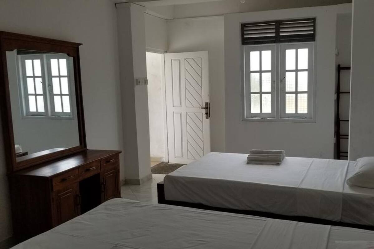 A bedroom with two beds, a dresser, a window with blinds, and a door leading to another room.