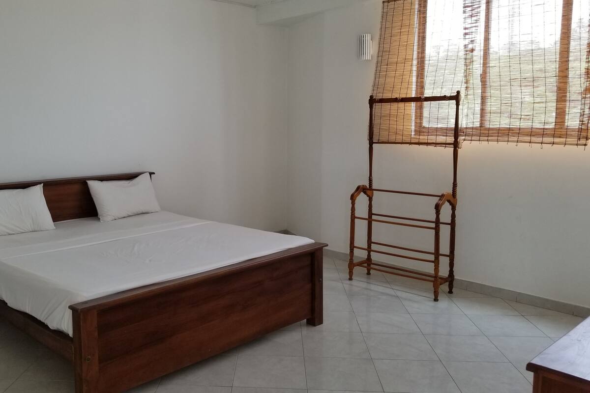 A bedroom with a wooden bed, white bedding, a wooden ladder, and a window with a bamboo blind.