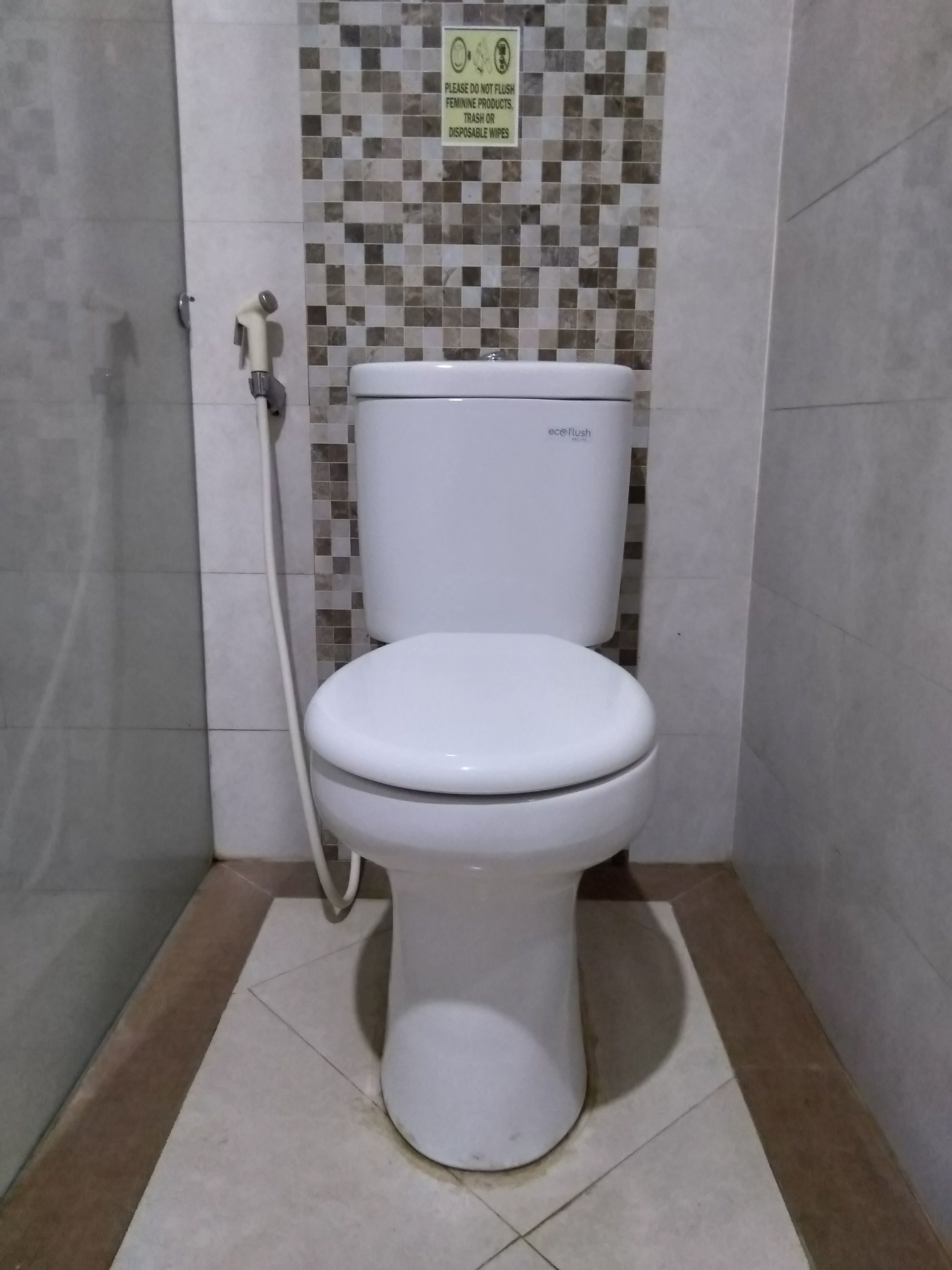 A white toilet with an attached bidet hose in a bathroom with tiled walls and floor.
