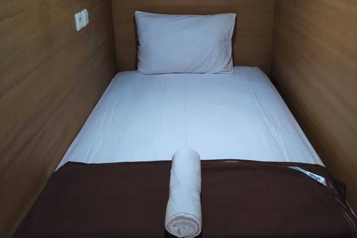 Una habitación pequeña con una cama individual, cabecera de madera, ropa de cama blanca y una toalla enrollada sobre la cama.