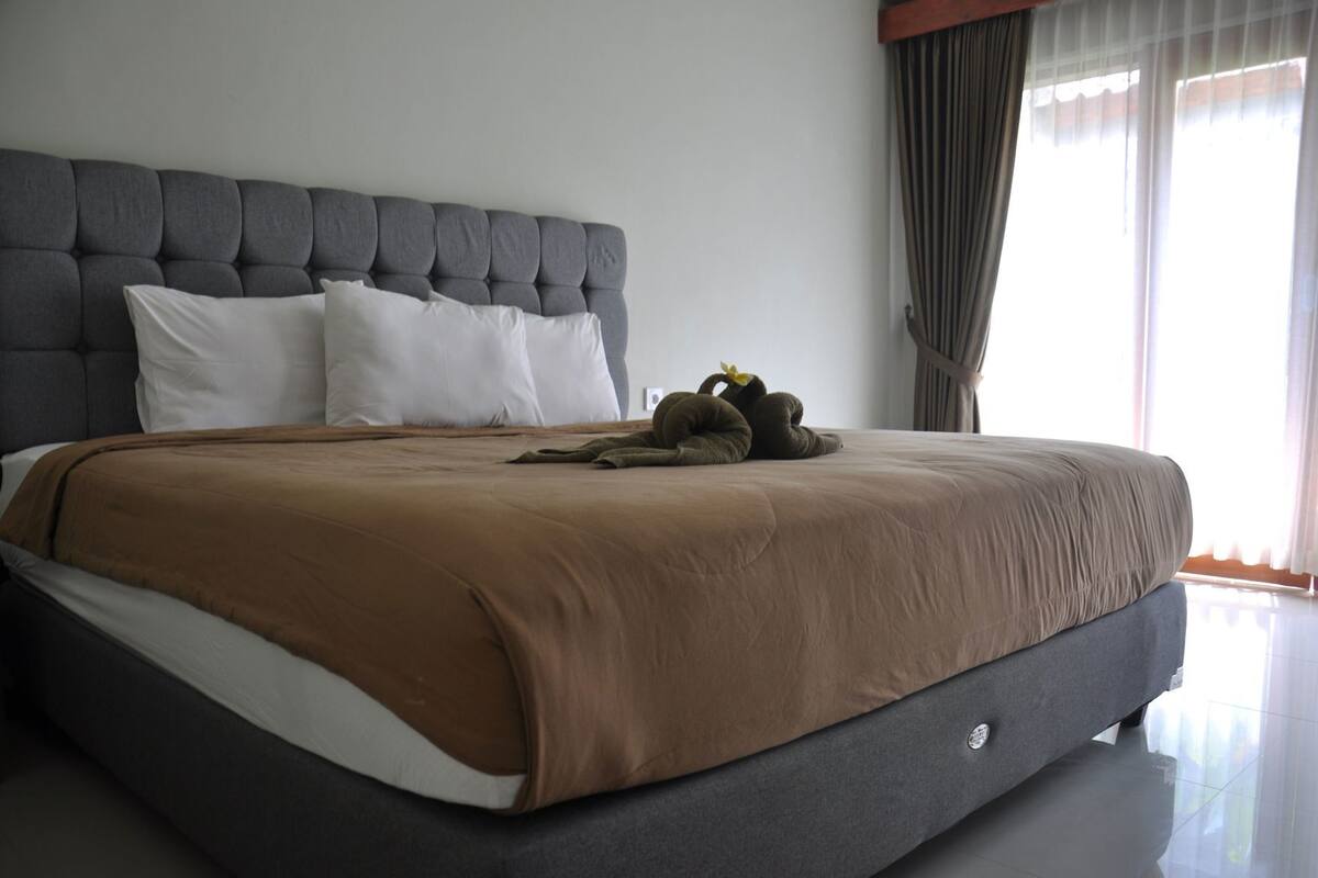 Una cama grande con cabecera gris, almohadas blancas y colcha marrón.
