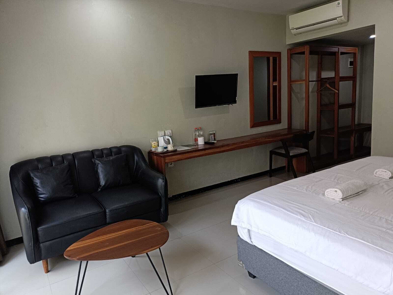 A hotel room with a black leather sofa, a wooden coffee table, a desk with a flat-screen TV, a bed with white bedding, and a wardrobe with a mirror.