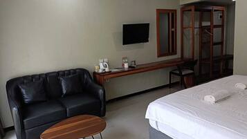 A hotel room with a black leather sofa, a wooden coffee table, a desk with a flat-screen TV, a bed with white bedding, and a wardrobe with a mirror.