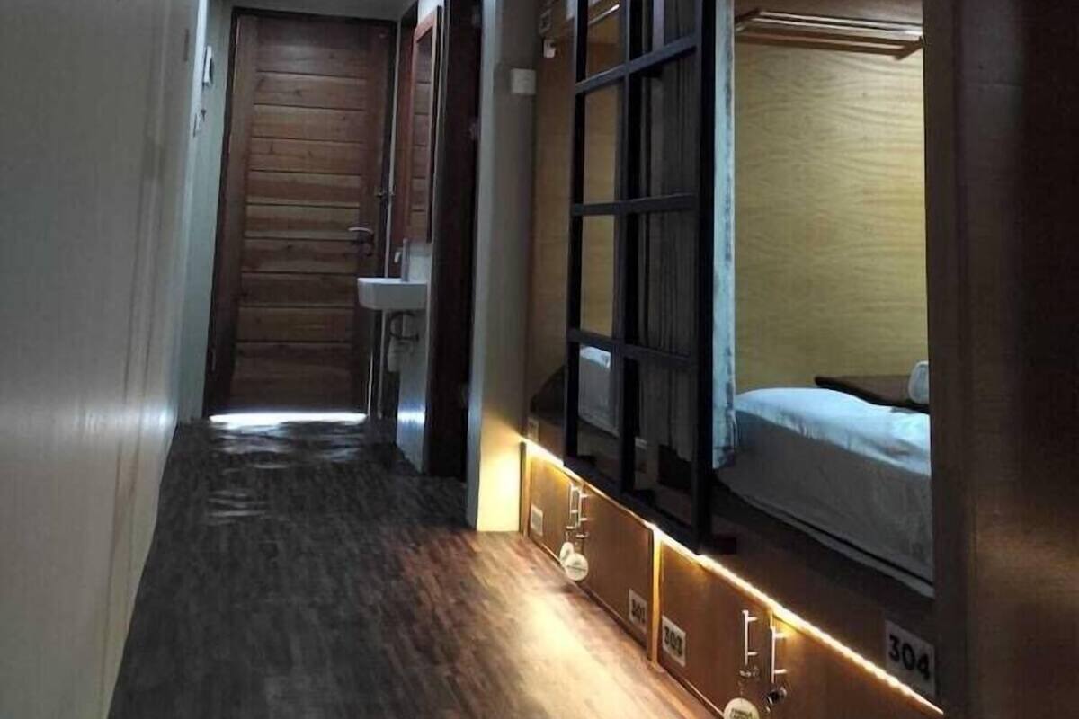 Un pasillo estrecho con suelo de madera, una puerta de madera al final y una cama litera con ventana a la derecha.