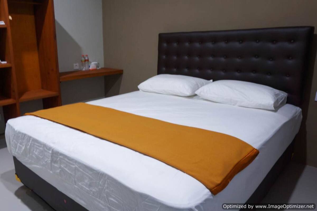 Una cama bien hecha con edredón blanco y una manta color mostaza, un cabecero tapizado oscuro y una mesita de noche de madera con un espejo y algunos objetos encima.