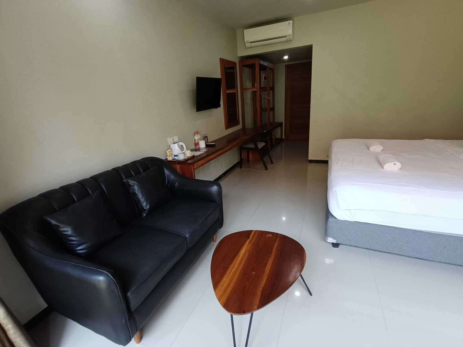 Una habitación de hotel con un sofá de cuero negro, una mesa de centro de madera, una cama con ropa blanca, un televisor montado en la pared y un equipo de aire acondicionado sobre la puerta.