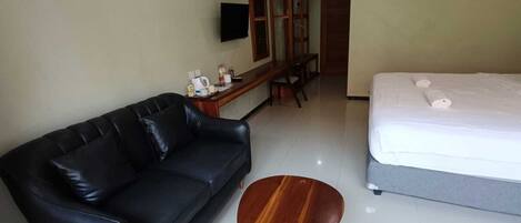 Una habitación de hotel con un sofá de cuero negro, una mesa de centro de madera, una cama con ropa blanca, un televisor montado en la pared y un equipo de aire acondicionado sobre la puerta.
