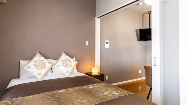 A bedroom with a bed, pillows, bedside table, lamp, and a TV mounted on the wall. - Mr.KINJO in Arcade (Naha)