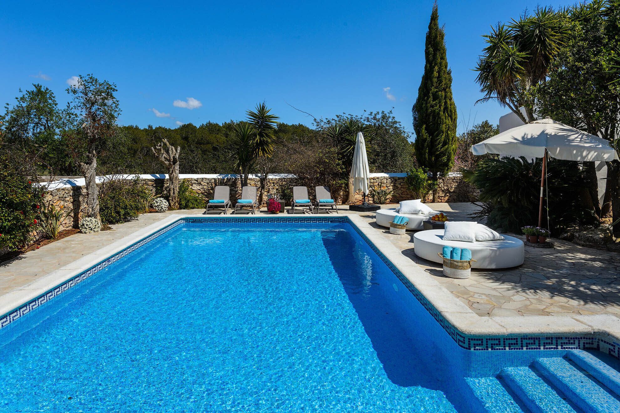 Una piscina rectangular con agua azul cristalina, rodeada de sillones de descanso y un muro de piedra.