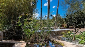 Un jardín tranquilo con un estanque, muros de piedra y plantas tropicales.