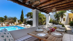 Zona de comedor junto a la piscina con mesa de madera para cuatro personas, rodeada de sillones de descanso y una pared blanca.