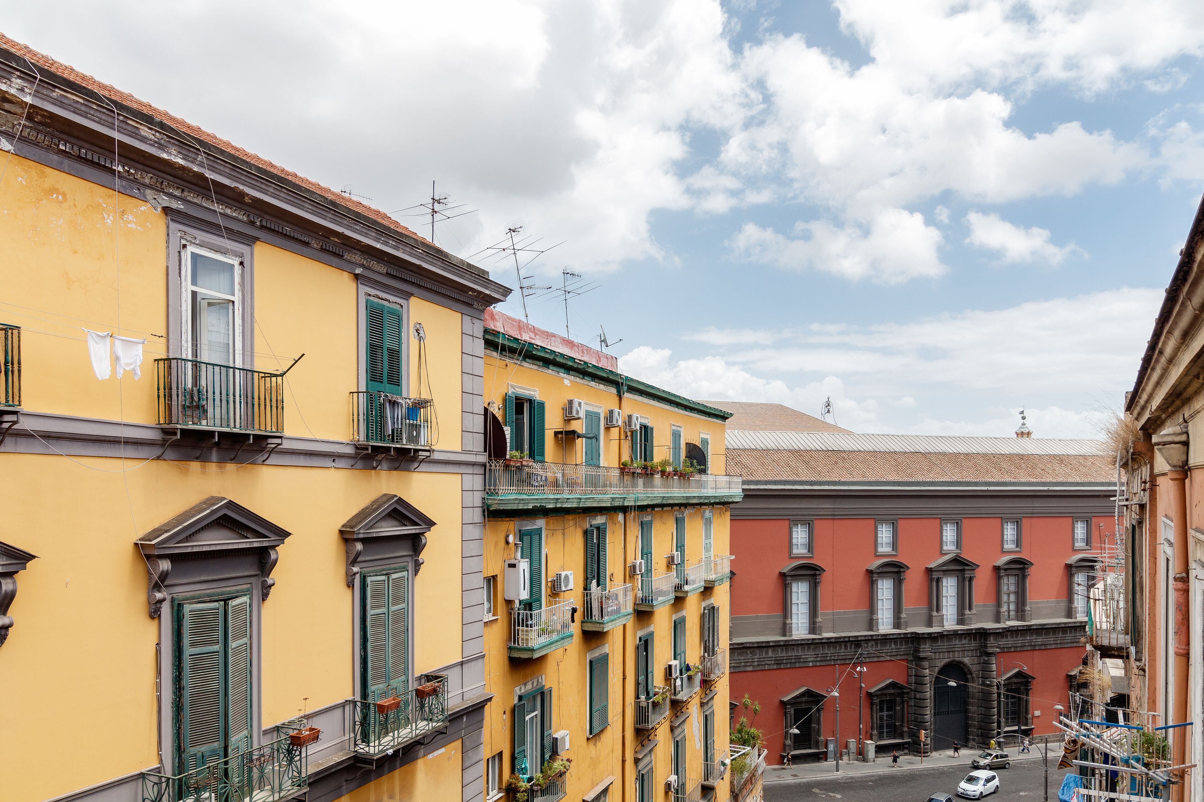 A row of yellow buildings with green shutters and balconies, a street in front, and a clear sky.
