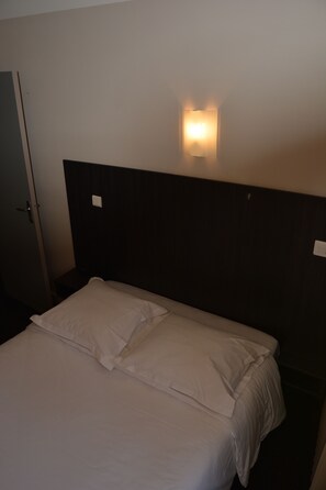A hotel room with a bed, a nightstand, and a wall-mounted light fixture. - Le Charleston Hotel (Rodez)