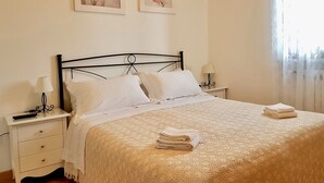 Een netjes opgemaakt bed met een gertextureerd deken, twee nachtkastjes met lampen en twee bloemrijke schilderijen aan de muur.
