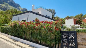 A modern two-story building with a white facade and a blue roof, surrounded by lush greenery and red flowers. - Luxury design villa with fantastic sea and mountain views (Betlem)