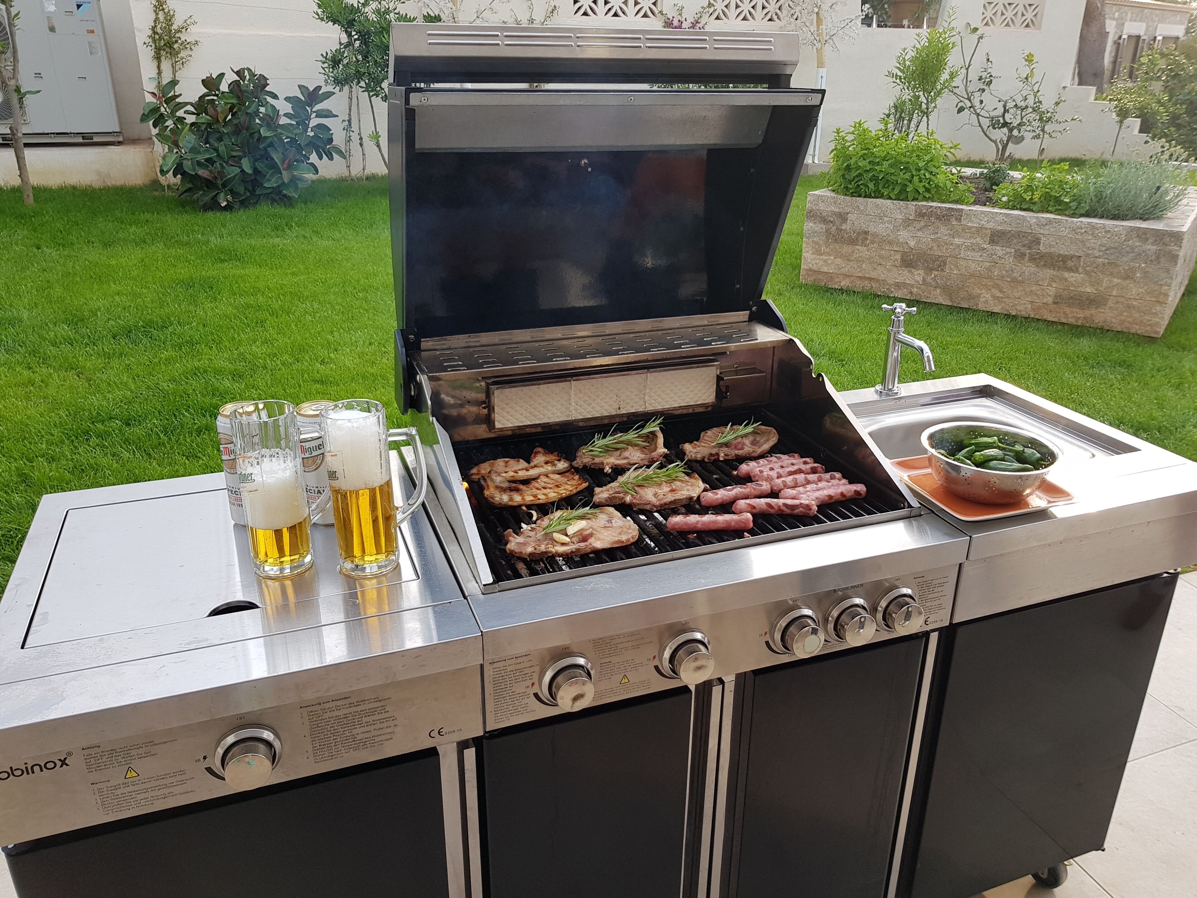 A portable grill with meat and sausages being cooked, a colander with vegetables, and two glasses of beer on a table.