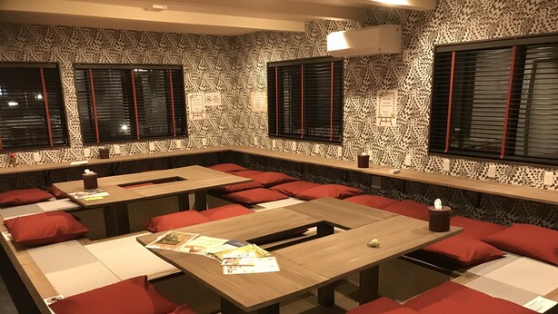 Ein modernes Restaurant mit langen Gemeinschaftstischen, roten Kissen, gemusterten Wänden und Fenstern mit Jalousien.