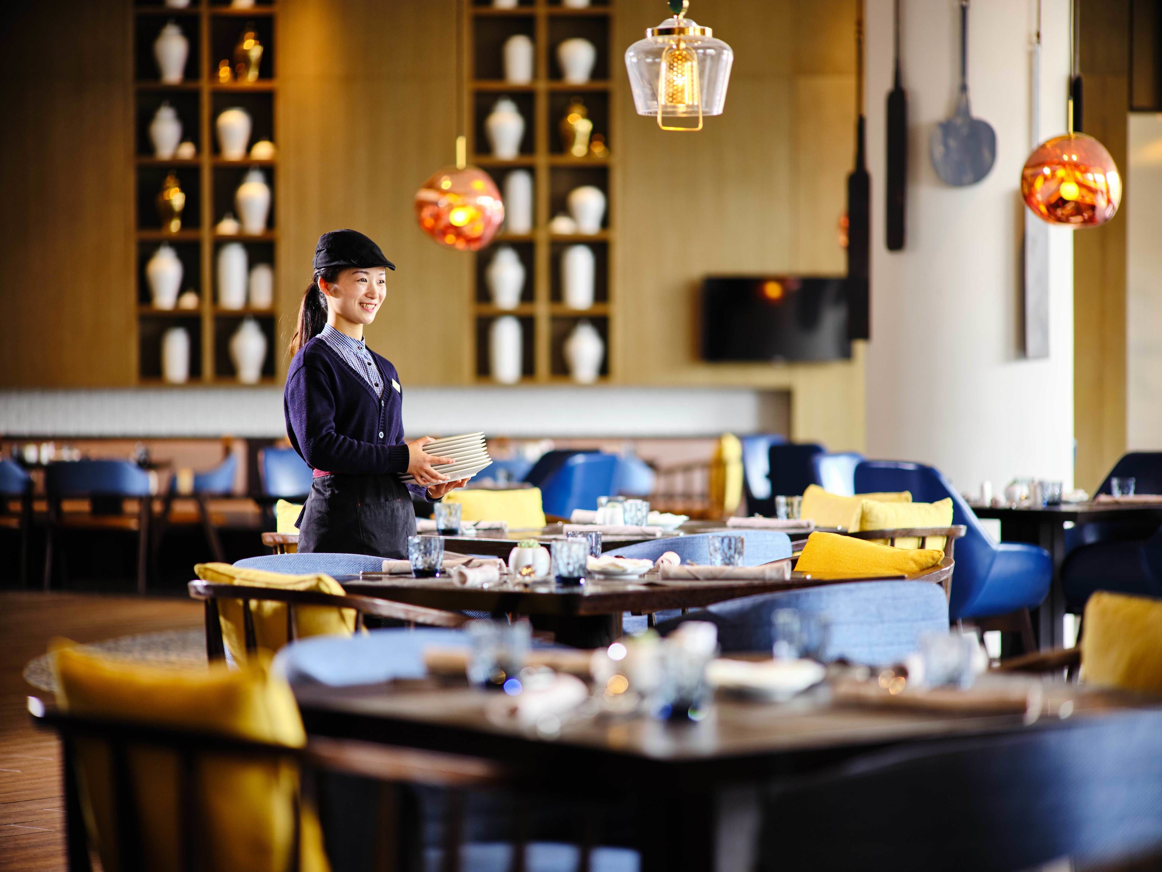Een restaurantinterieur met gedekte tafels, een ober met een stapel borden, en hangende lantaarns.