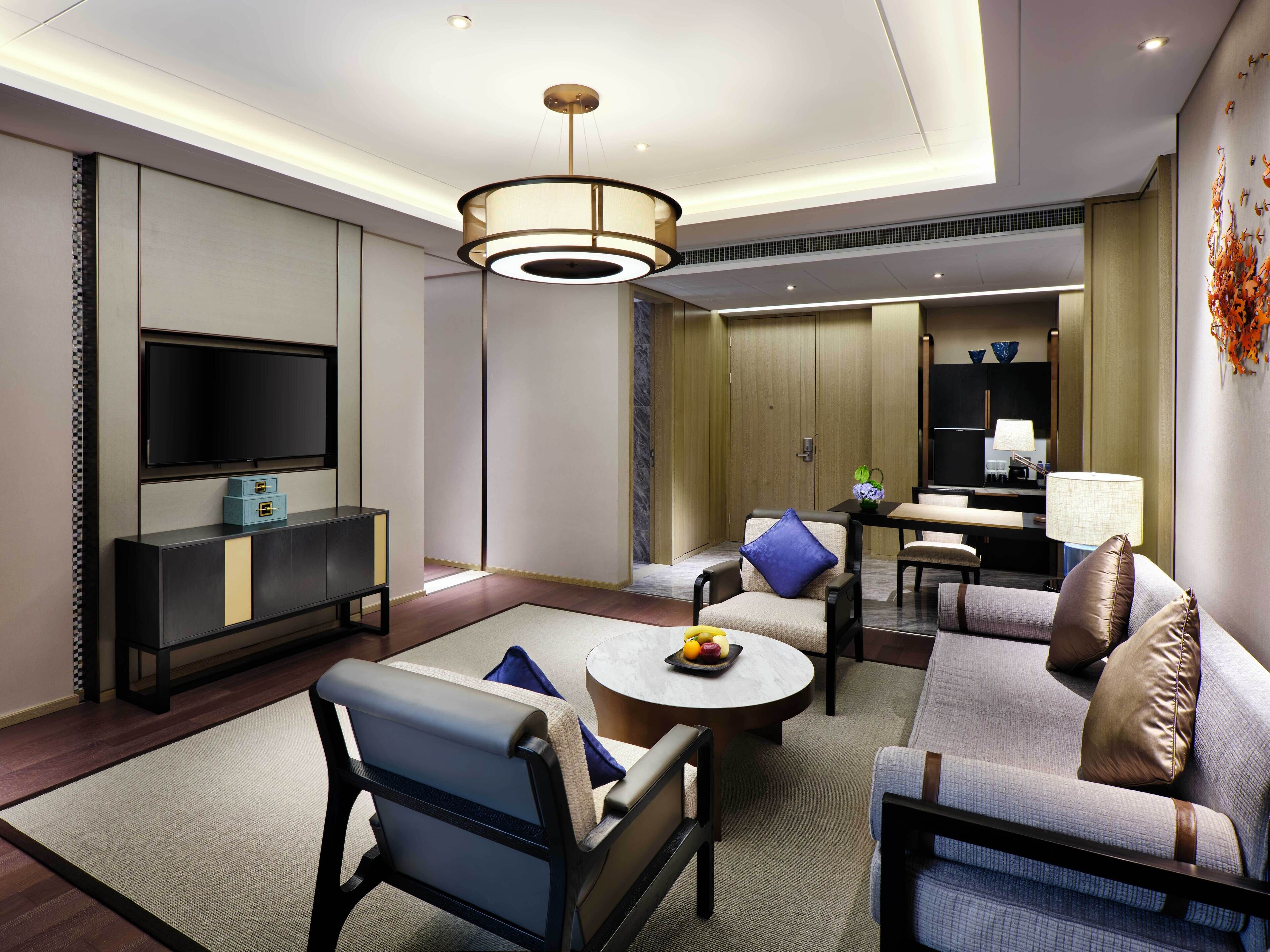 A modern hotel room with a sofa set, a TV, a dining table, and a minibar.