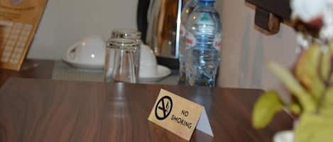 A hotel room with a wooden table, a no smoking sign, a water bottle, a coffee maker, and a telephone.