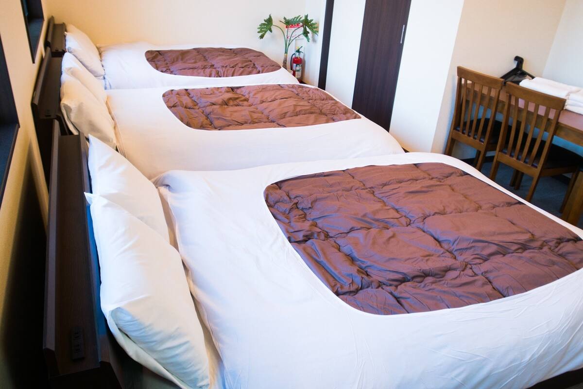 Een tweepersoonsbed met een bruine quilt en witte kussens, een nachtkastje van hout met een plant en een houten stoel in een hotelkamer.