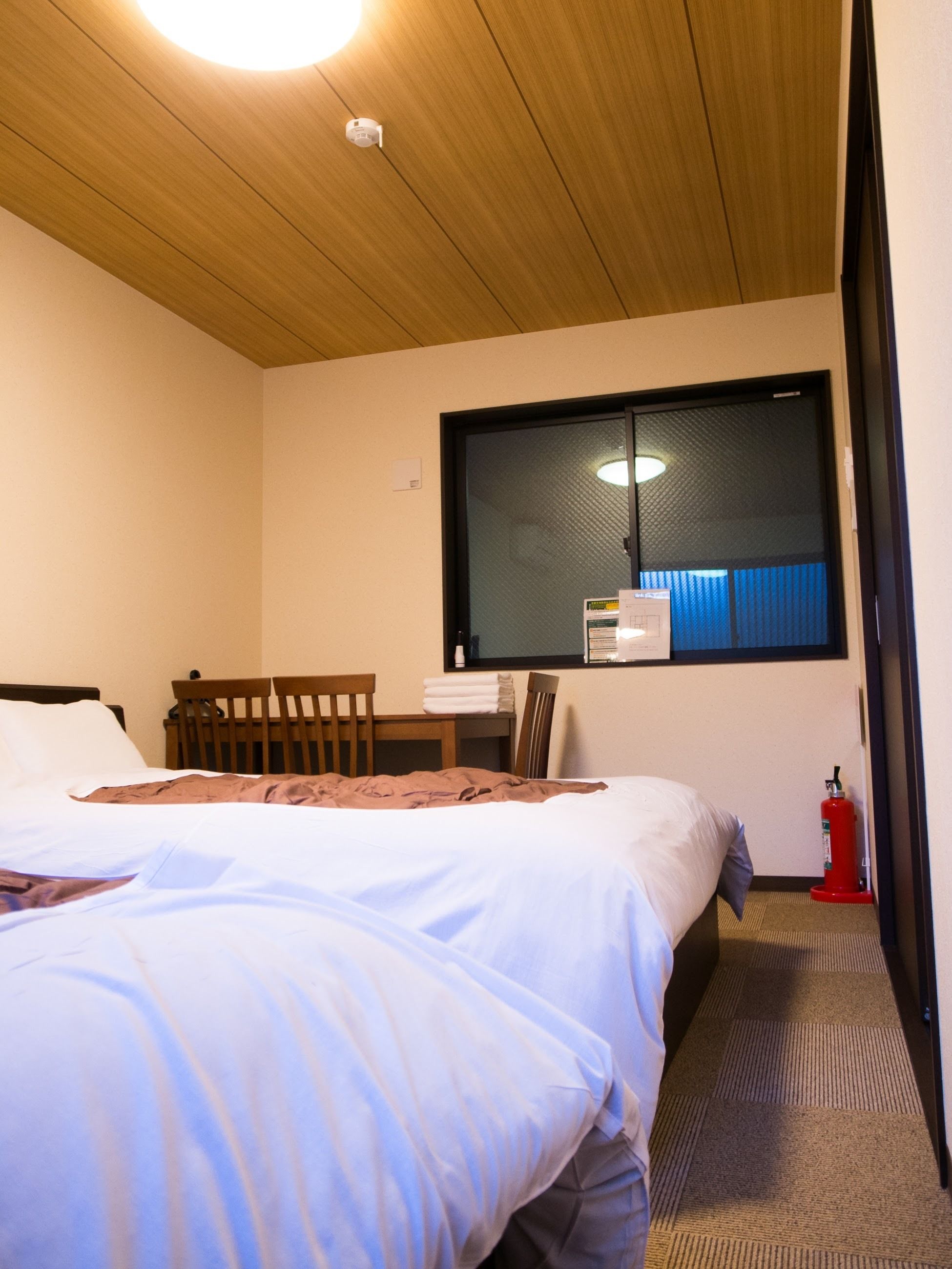 Ett hotellrum med ett träpanelat tak, en stor säng med vita sängkläder, ett skrivbord med en stol, ett fönster med persienner och en röd brandsläckare.