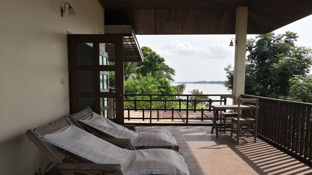 A balcony with two wooden lounge chairs, a table, and a view of a body of water. - Keerawan House Rim Khong (Phon Phisai)